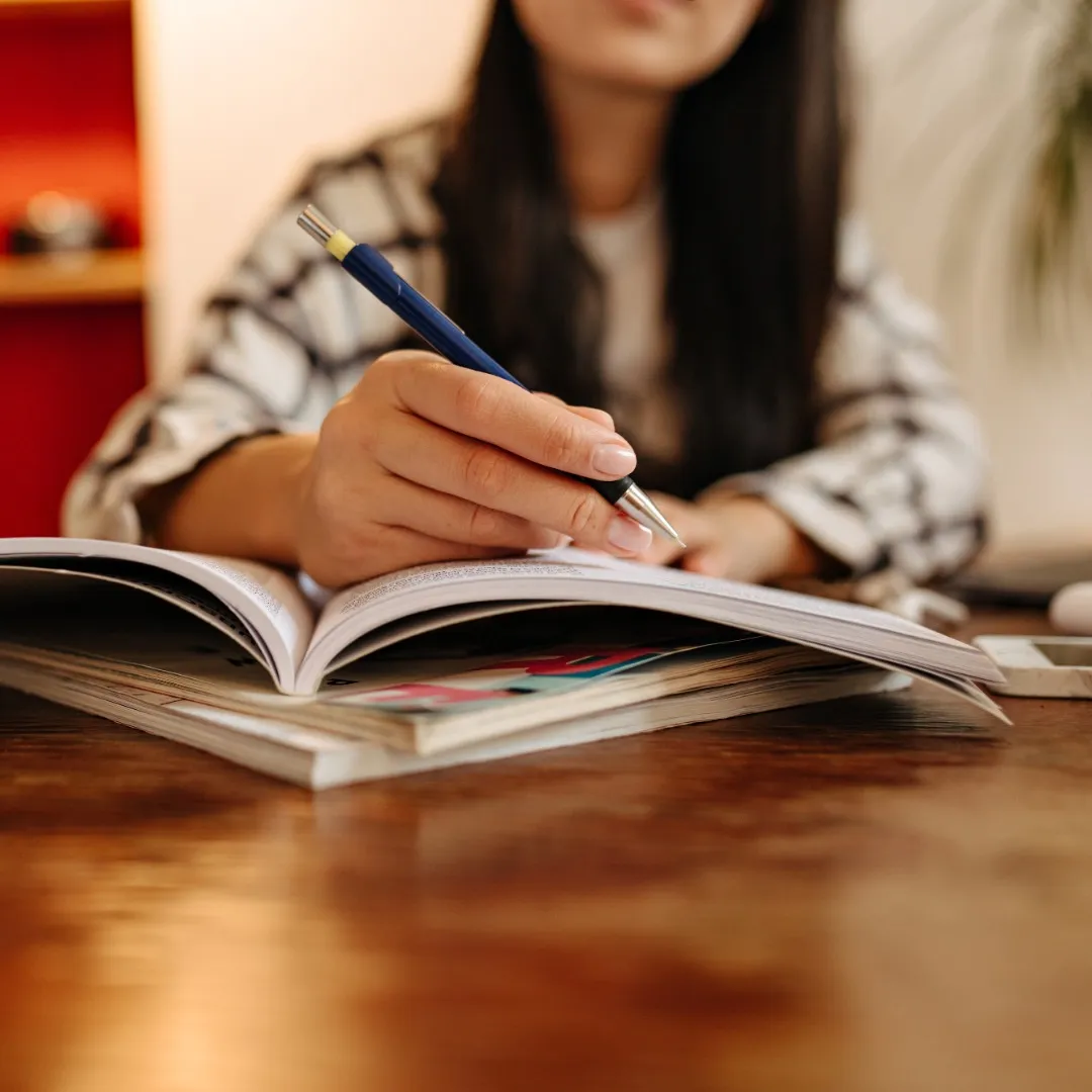 Une personne concentrée engagée dans l'écriture ou l'étude, avec un stylo à la main sur un cahier ouvert sur un bureau en bois.