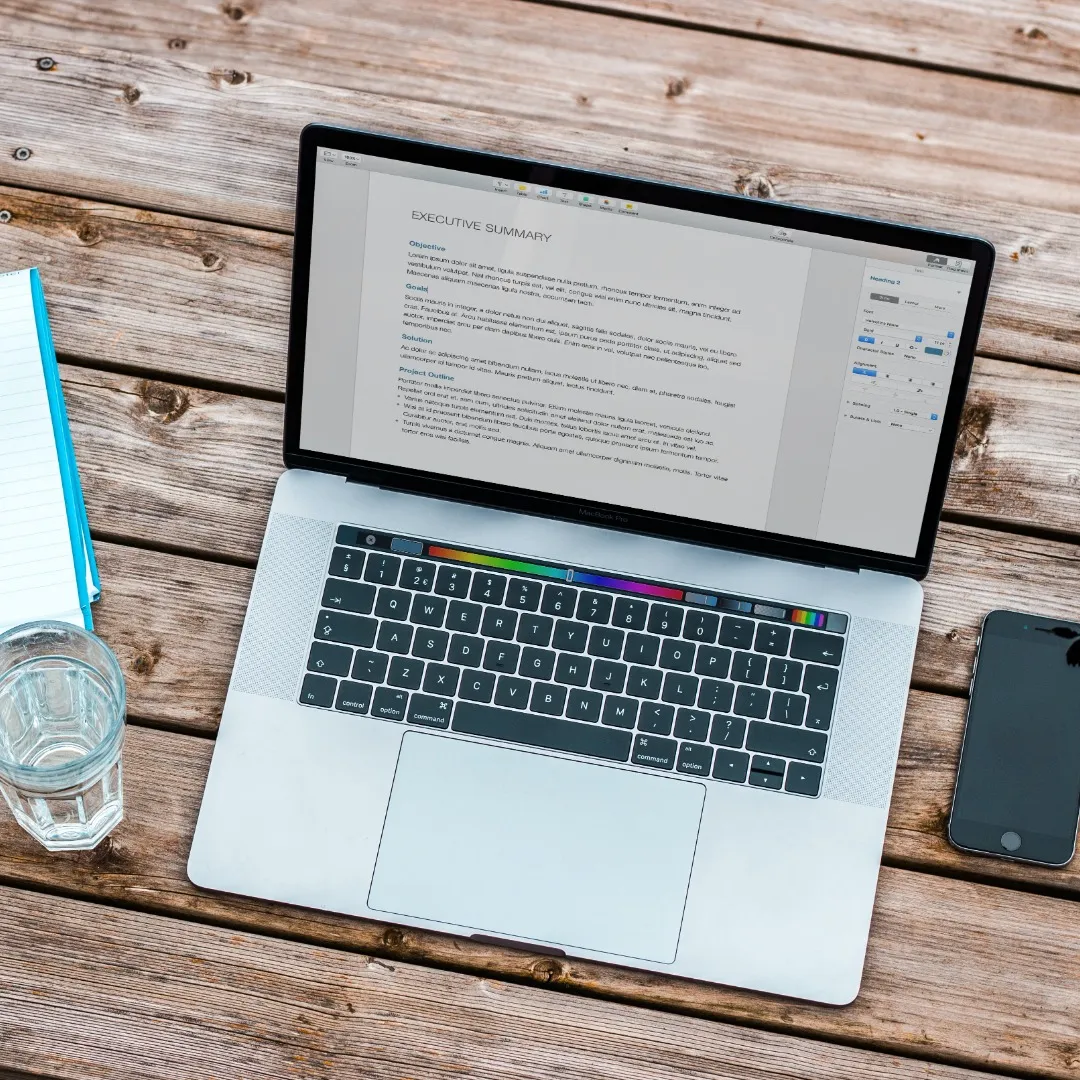 Un ordinateur portable moderne affichant un document de synthèse, accompagné d'un smartphone, d'un verre d'eau et de papier à lettres, soigneusement disposés sur une surface en bois.